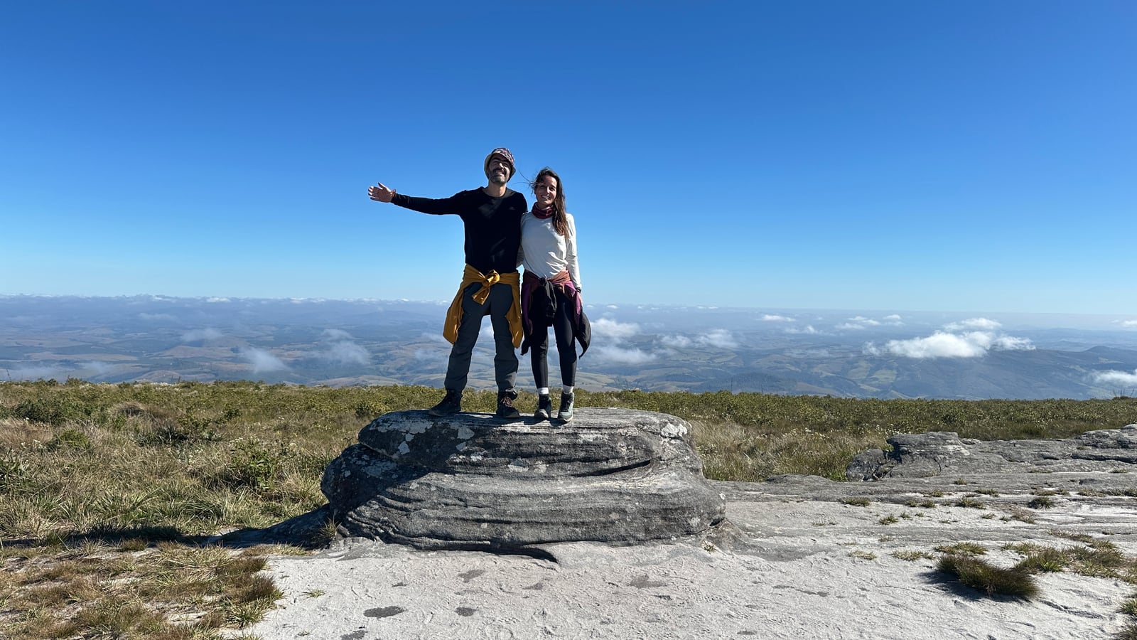 Thaíssa e Julián de pé numa pedra alta, de braços abertos, com o vale atrás