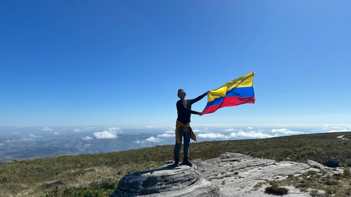Julián de pé numa pedra, acima das nuvens, segurando a bandeira da Colômbia