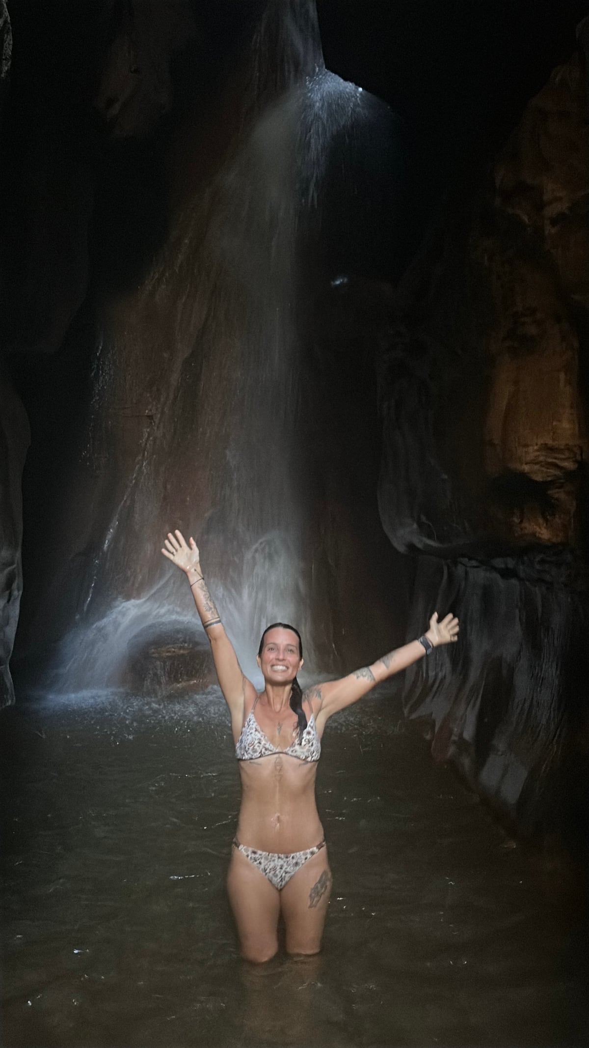 Thaíssa de braços abertos debaixo de uma cachoeira, dentro de uma gruta de pedra